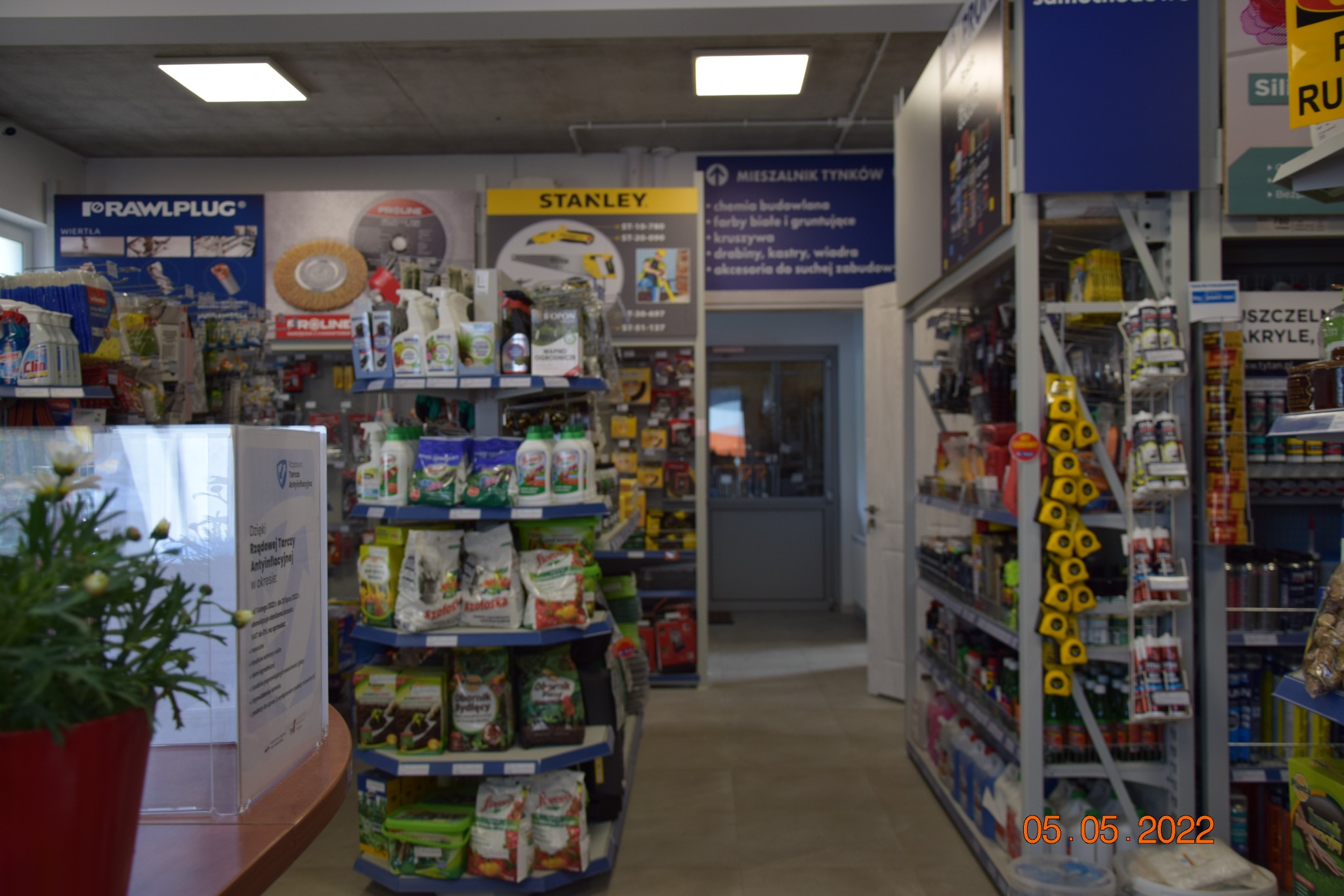 Wnętrze sklepu budowlanego z regałami pełnymi materiałów i narzędzi, widoczne reklamy marek Rawlplug i Stanley, oraz tablica informacyjna Mieszalnik Tynków.
