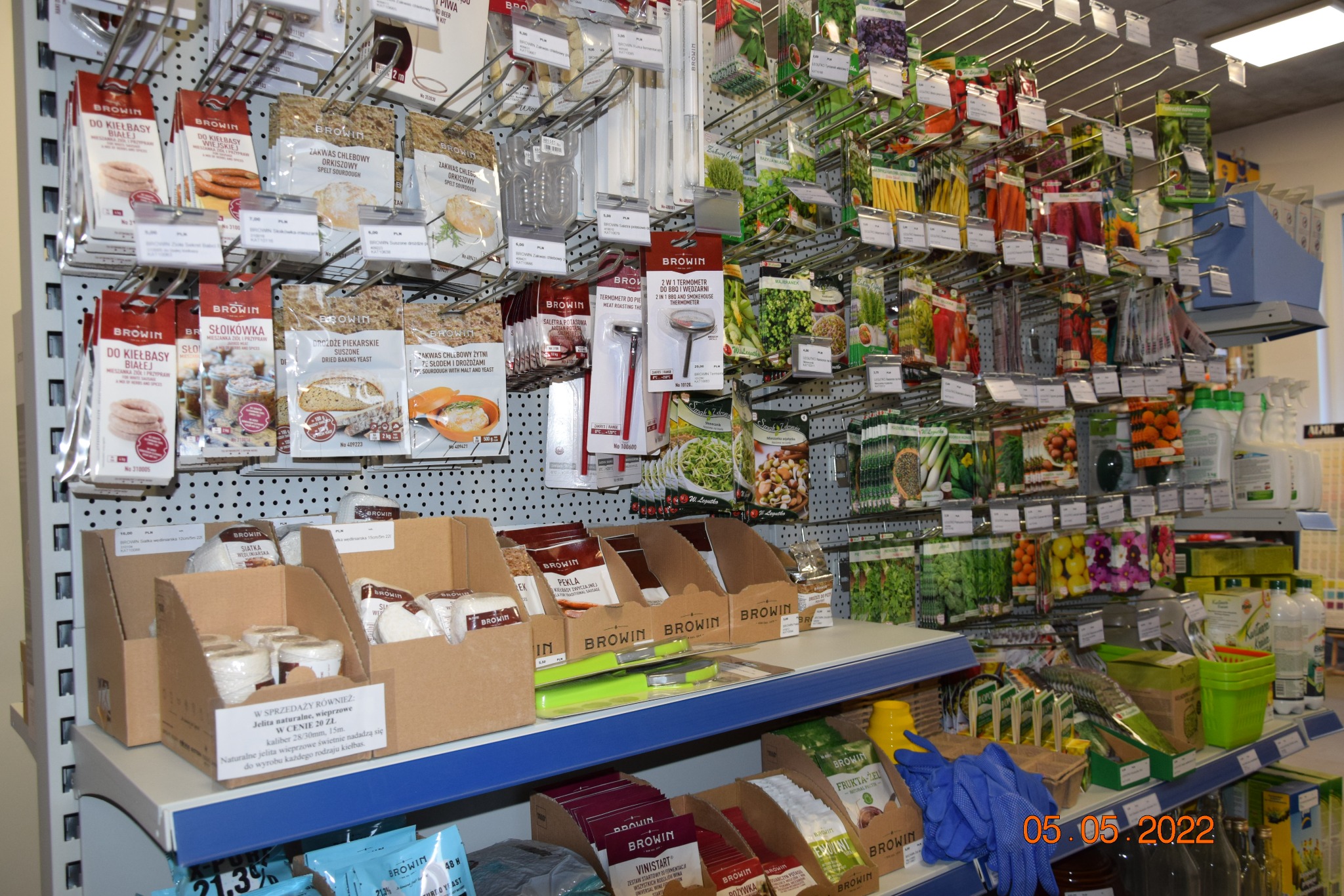 Półka sklepowa z artykułami do wyrobu wędlin i przetworów domowych: przyprawy, jelita, drożdże, termometry, nasiona, środki czyszczące.