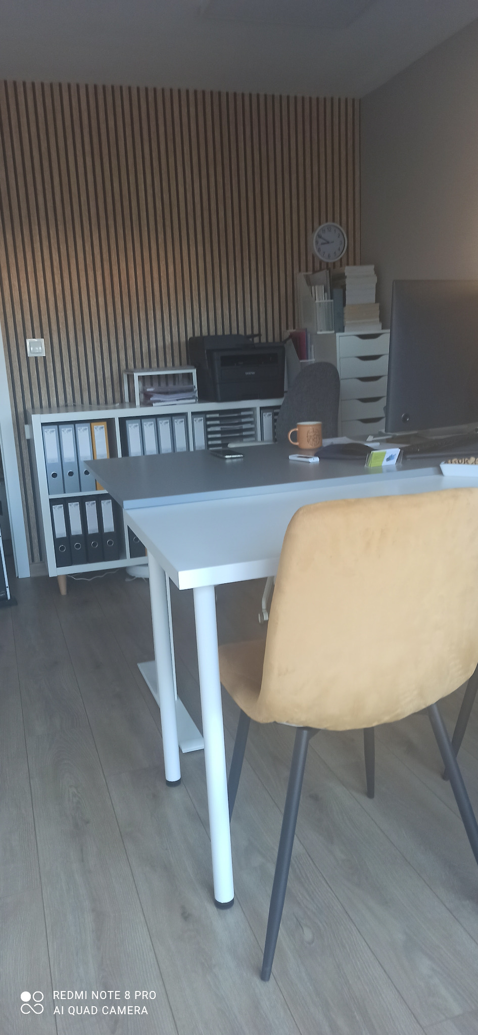 Nowoczesne biuro z drewnianą ścianą, białe regały z segregatorami i drukarką, biurko z monitorem i kubkiem. Krzesło w kolorze musztardowym. Stonowana kolorystyka.