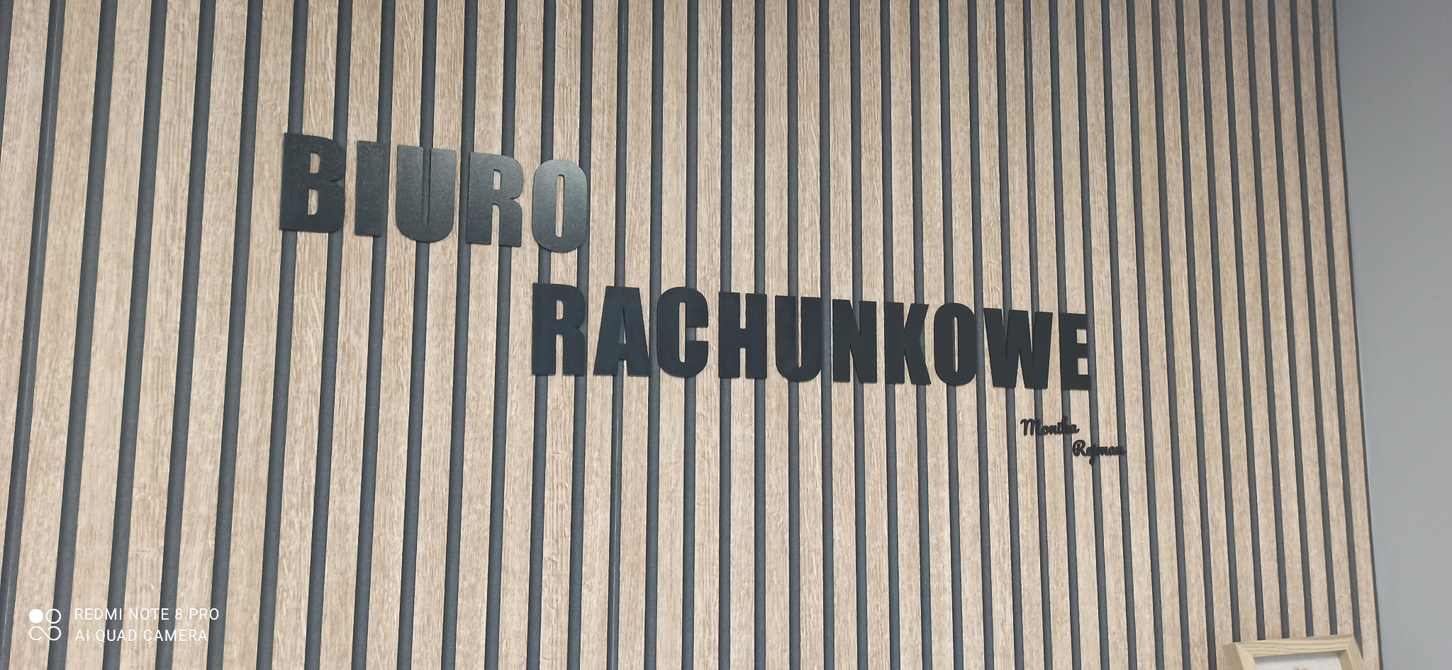 Ściana z drewnianych paneli z napisem 'Biuro Rachunkowe' w kolorze czarnym i podpisem 'Monika Rejman'.