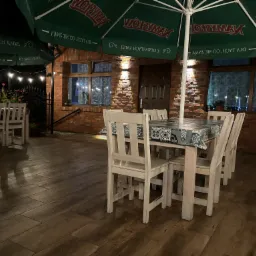 Zewnętrzny taras restauracji z drewnianą podłogą, stołem nakrytym w niebiesko-białą kratę i białymi krzesłami, oświetlony lampkami i parasolami z logo, na tle ceglanego budynku.