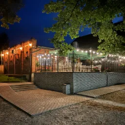 Oświetlony budynek restauracji z ogródkiem w wieczornej scenerii, widoczne drzewa, ceglana fasada i dekoracyjne lampki