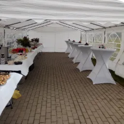 Wnętrze białego namiotu cateringowego z dwiema długimi zastawionymi stołami i rzędem mniejszych stolików koktajlowych z piramidami kieliszków.