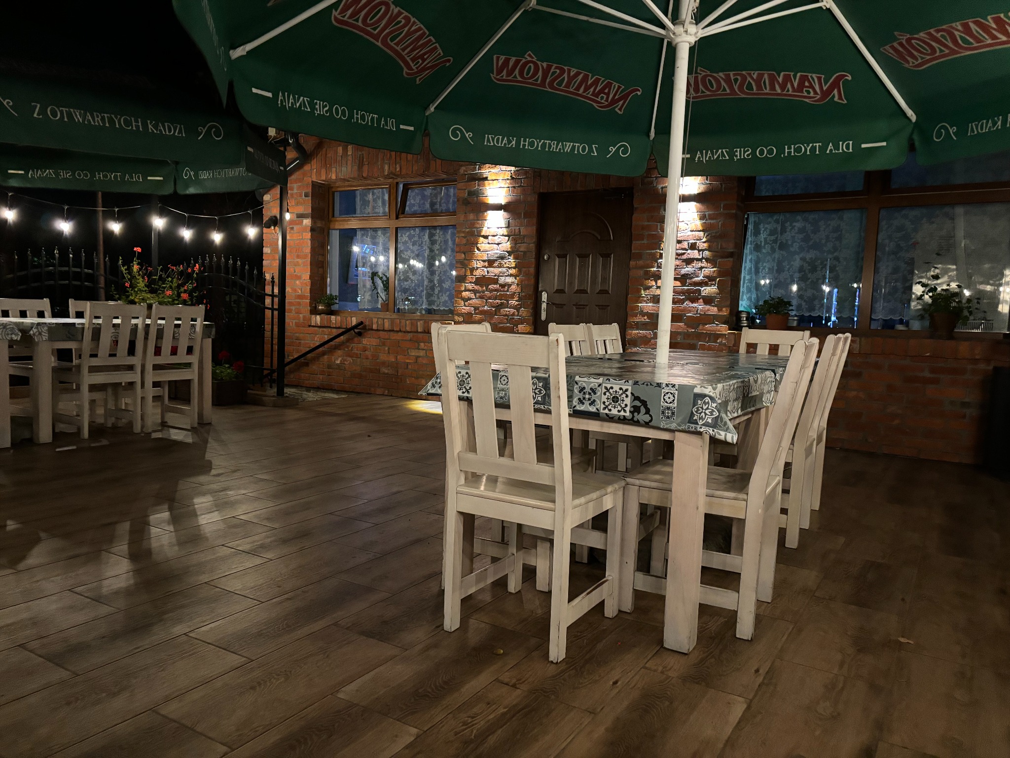 Zewnętrzny taras restauracji z drewnianą podłogą, stołem nakrytym w niebiesko-białą kratę i białymi krzesłami, oświetlony lampkami i parasolami z logo, na tle ceglanego budynku.