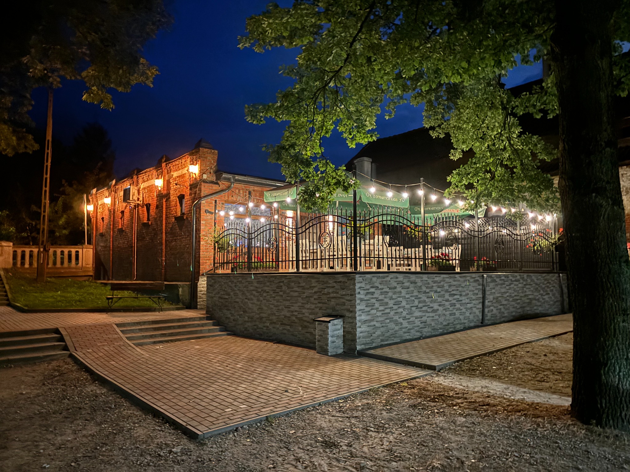 Oświetlony budynek restauracji z ogródkiem w wieczornej scenerii, widoczne drzewa, ceglana fasada i dekoracyjne lampki