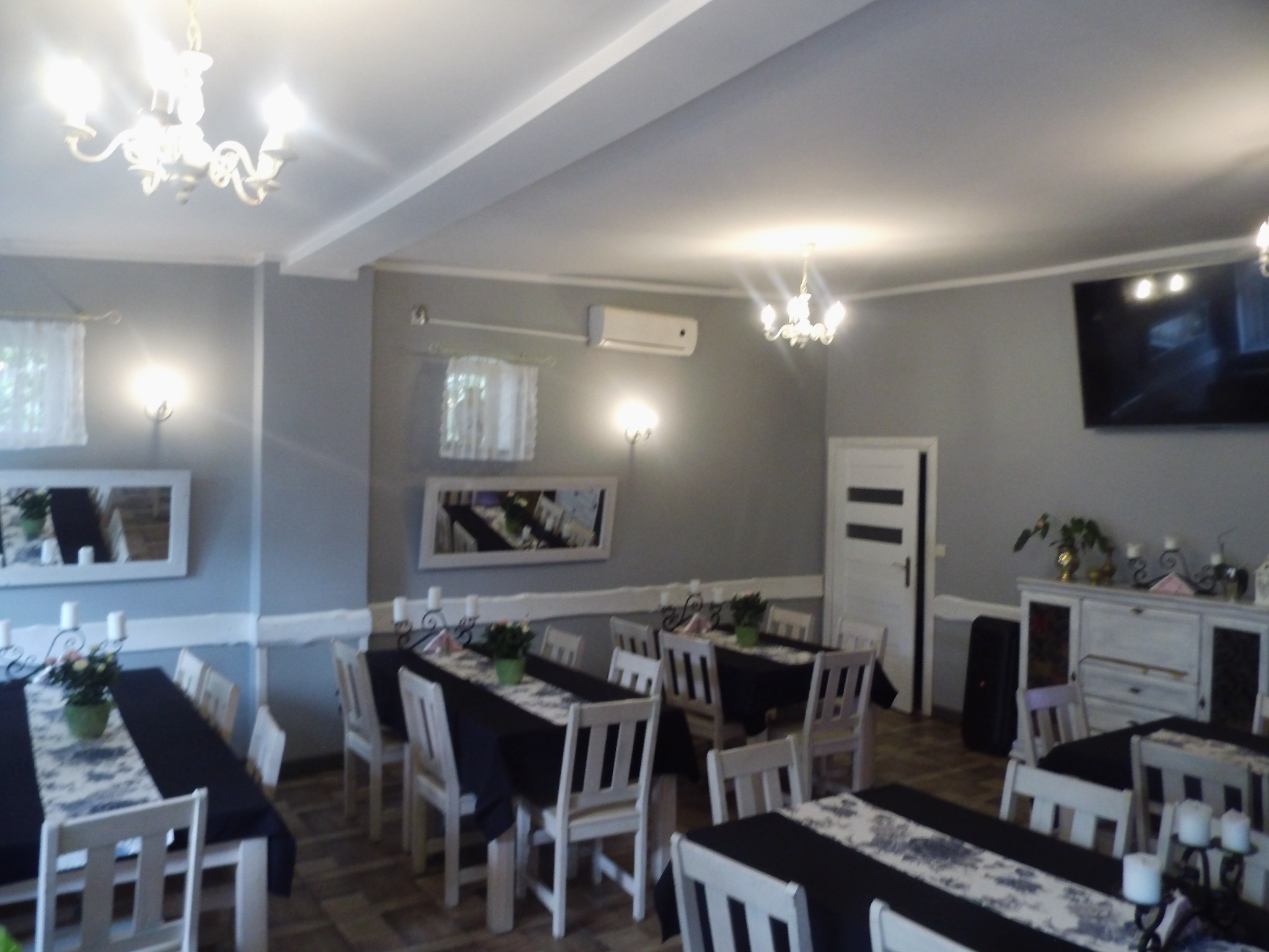 Sala bankietowa z ustawionymi stołami, nakrytymi czarnymi obrusami i białymi bieżnikami w kwiatowy wzór. Białe krzesła, świeczniki ze świecami i małe bukiety kwiatów na stołach. Na ścianach lustra...