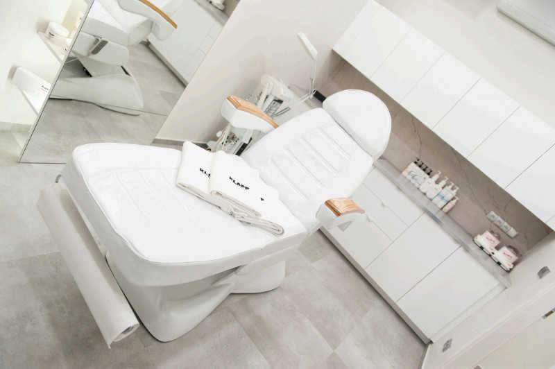 Biały, skórzany fotel kosmetyczny z drewnianymi podłokietnikami i ręcznikami Klapp w nowoczesnym, minimalistycznym salonie. W tle widoczne białe szafki i półki z kosmetykami.