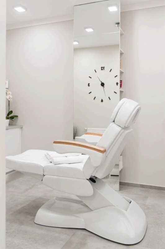 Biały, regulowany fotel kosmetyczny z drewnianymi podłokietnikami w minimalistycznym, jasnym wnętrzu salonu z zegarem ściennym i lustrem.