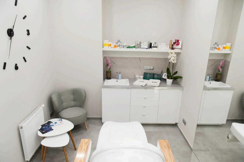 Jasne wnętrze gabinetu kosmetycznego z białymi meblami, umywalką, dekoracjami kwiatowymi i zegarem na ścianie, fotel zabiegowy na pierwszym planie.