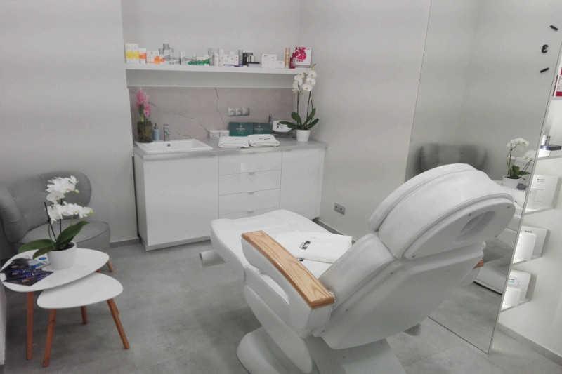 Jasne wnętrze gabinetu kosmetycznego z fotelem zabiegowym, umywalką, szafkami z produktami i dekoracyjnymi orchideami.