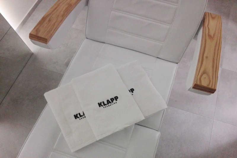 Biały fotel kosmetyczny z drewnianymi podłokietnikami i złożonymi ręcznikami z logo Klapp Cosmetics.