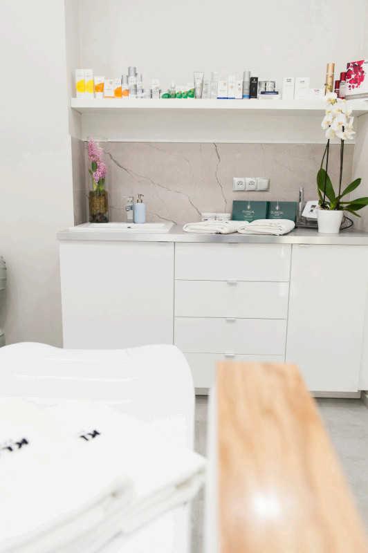 Jasne wnętrze salonu kosmetycznego: białe meble, kosmetyki na półce, kwiaty w wazonie i doniczce, leżanka kosmetyczna z ręcznikami z logo.