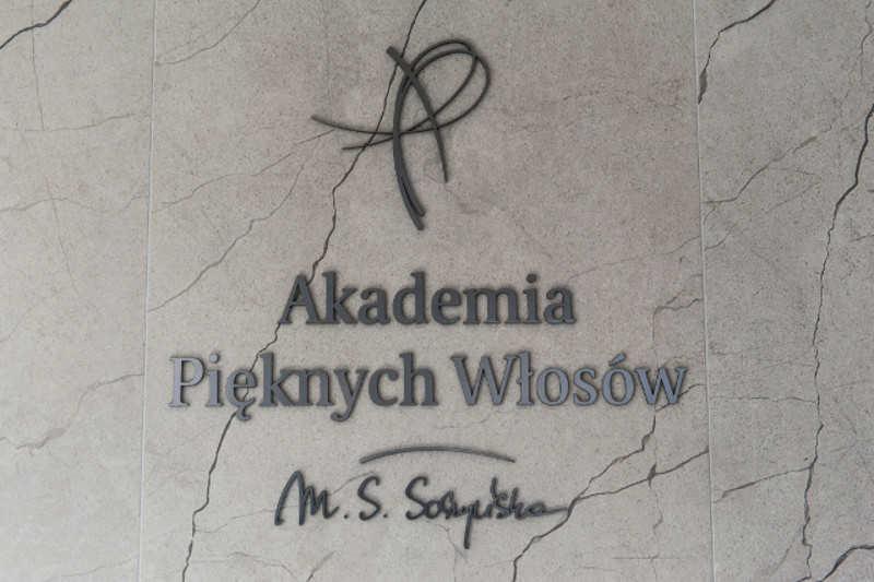 Logo Akademii Pięknych Włosów z abstrakcyjnym symbolem nad nazwą, umieszczone na marmurowej ścianie z widocznymi żyłkami.