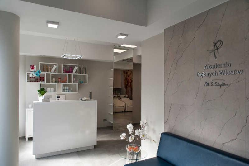 Recepcja w jasnym salonie z logo 'Akademia Pięknych Włosów' na marmurowej ścianie, białe regały z kosmetykami i kwiatami, minimalistyczny wystrój.