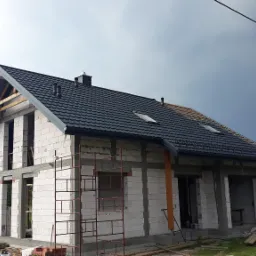 Dom w trakcie budowy z częściowo położoną blachodachówką modułową w kolorze grafitowym; widoczna więźba dachowa, ściany z betonu komórkowego oraz rusztowanie.