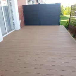 Jasny, kompozytowy taras przydomowy z desek w odcieniach beżu, z widoczną ścianą z cegły i ciemnym panelem ogrodzeniowym. Ujęcie z poziomu tarasu.
