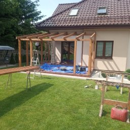 FPHU Daszko Vitalii Solovei - Montaż drewnianej konstrukcji pergoli przy domu jednorodzinnym. Na trawniku leżą narzędzia i materiały budowlane. Prace w toku, widoczny pracownik na drabinie.