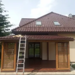 Drewniana pergola z żaluzjami wbudowana w bryłę domu, widoczna drabina, brązowa dachówka, zielone drzewa w tle.