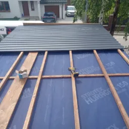 Montaż zadaszenia: drewniany szkielet konstrukcji pokryty folią, częściowo przykryty szarą blachą trapezową. W tle dom z garażem i zaparkowane auta.