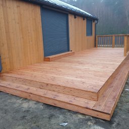 FPHU Daszko Vitalii Solovei - Nowy taras z drewna na zewnątrz budynku z drewnianą elewacją i szarą roletą. Widoczne schody wejściowe i balustrada. Całość w naturalnej kolorystyce drewna.