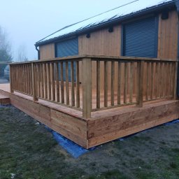 FPHU Daszko Vitalii Solovei - Drewniany taras z balustradą przy domu z drewnianą elewacją i szarymi roletami. Widoczne schody wejściowe. Mglista pogoda.