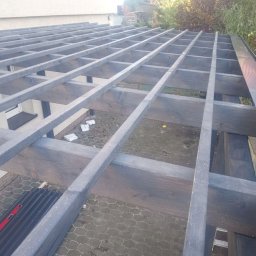 FPHU Daszko Vitalii Solovei - Konstrukcja drewnianej pergoli w trakcie budowy, widok z góry. Szare, pomalowane belki tworzą regularną siatkę nad tarasem z kostki brukowej. Widoczny poziomica.