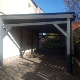 FPHU Daszko Vitalii Solovei - Ciemnoszara, drewniana pergola przy białym budynku, z widocznym brukowanym podjazdem i fragmentem ogrodu w tle. Konstrukcja oświetlona promieniami słońca.