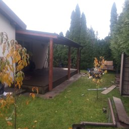 FPHU Daszko Vitalii Solovei - Zadaszony taras z drewnianą podłogą i brązowymi słupami, widok z ogrodu. W tle wysokie tuje i fragment domu. Na pierwszym planie drzewko z żółtymi liśćmi.