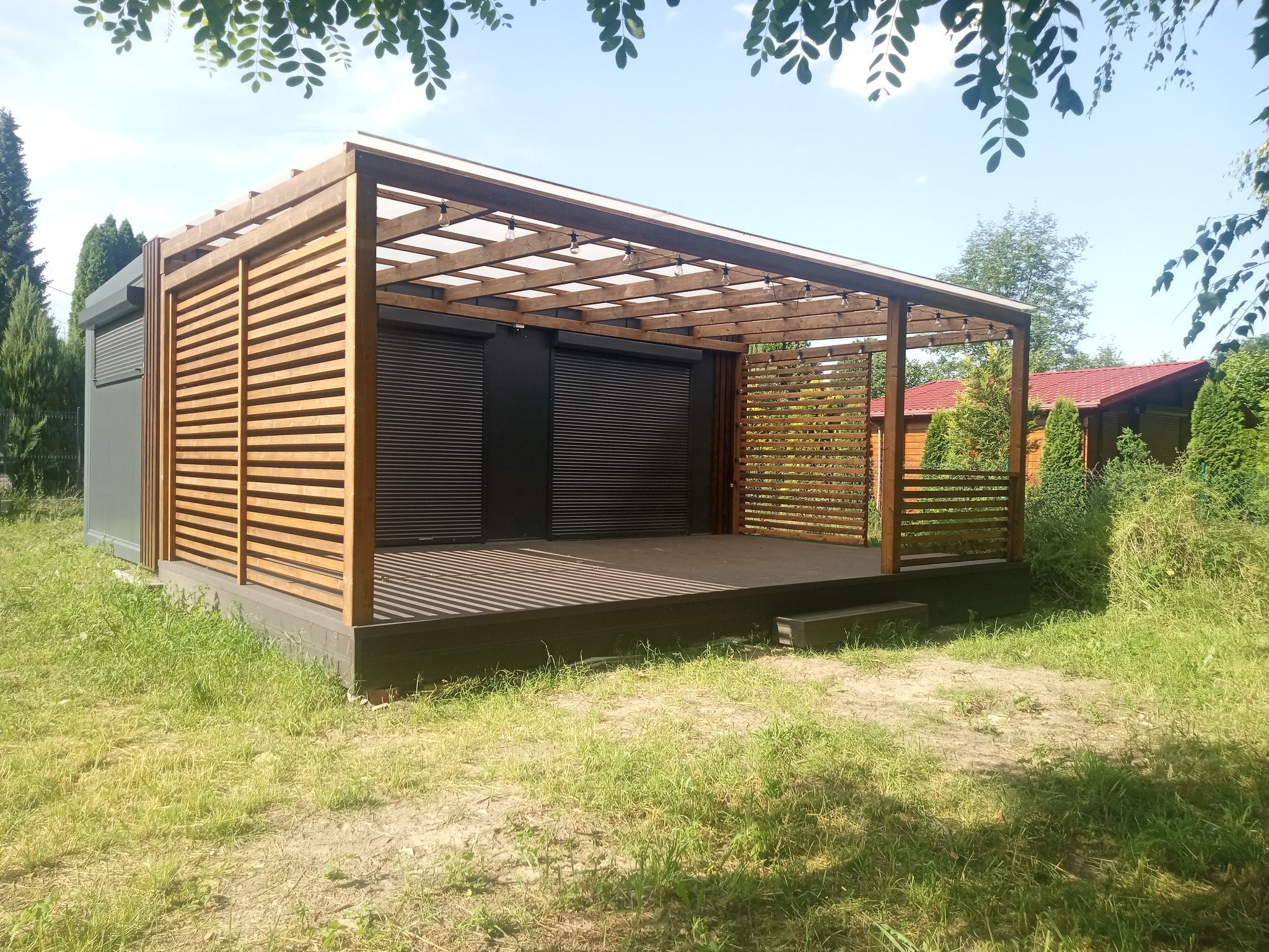 Drewniana pergola z zadaszeniem i roletami, oświetlona sznurem żarówek, otoczona zielenią. Widoczne elementy konstrukcyjne i wykończeniowe w naturalnym odcieniu drewna.