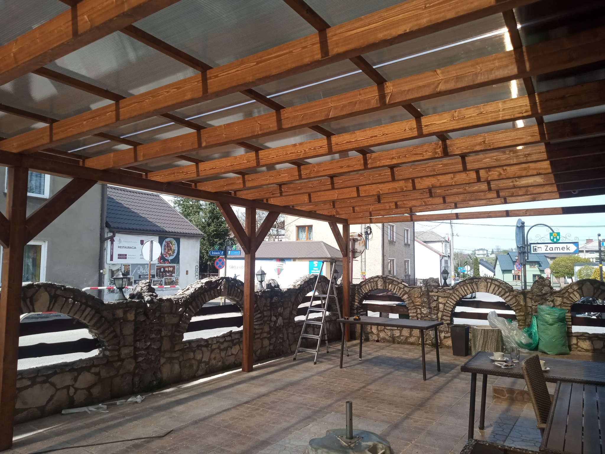 Zadaszona drewniana konstrukcja z przezroczystym dachem, wsparta na kamiennym murku z łukami. Widoczne stoły i krzesła, sugerujące przestrzeń restauracyjną.
