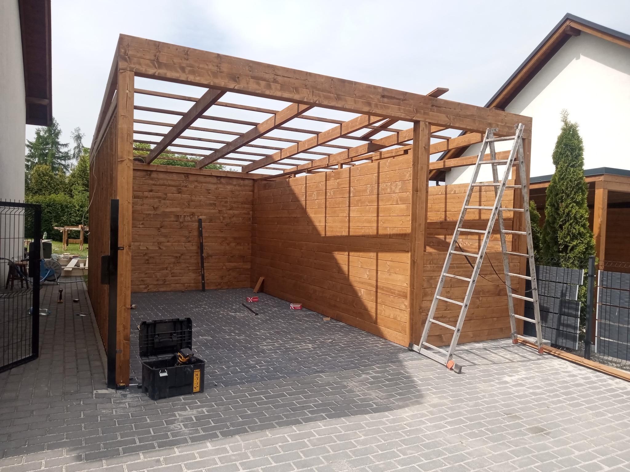 Drewniana konstrukcja z drabiny oparta o ścianę, skrzynka narzędziowa na szarym bruku. Widok na budowę drewnianej pergoli w ogrodzie.