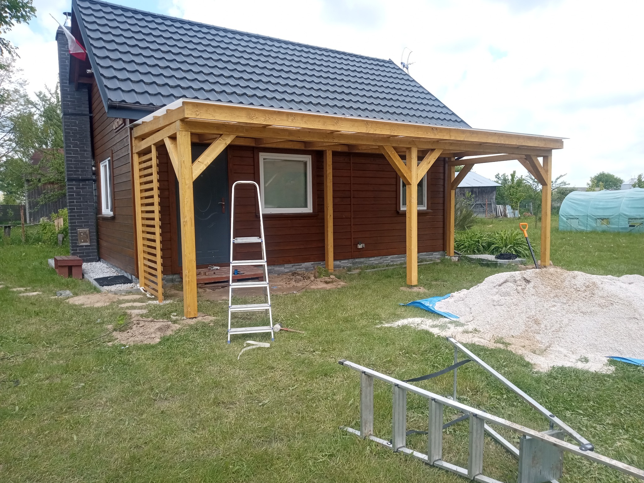 Drewniana pergola z przezroczystym zadaszeniem przy brązowym domu z szarą dachówką. Na trawniku drabiny i piasek. Widoczne elementy konstrukcyjne i wykończeniowe.
