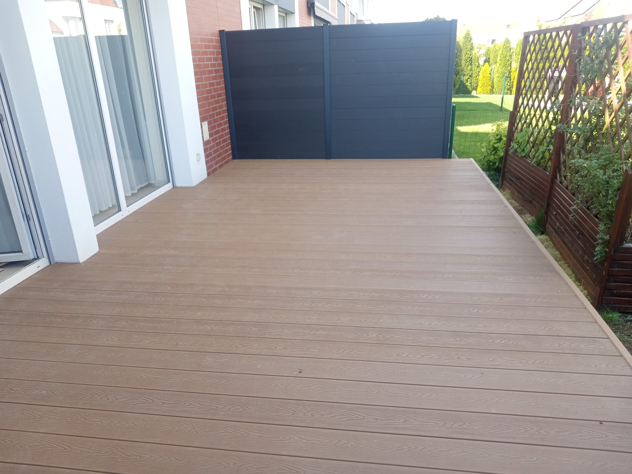 Jasny, kompozytowy taras przydomowy z desek w odcieniach beżu, z widoczną ścianą z cegły i ciemnym panelem ogrodzeniowym. Ujęcie z poziomu tarasu.