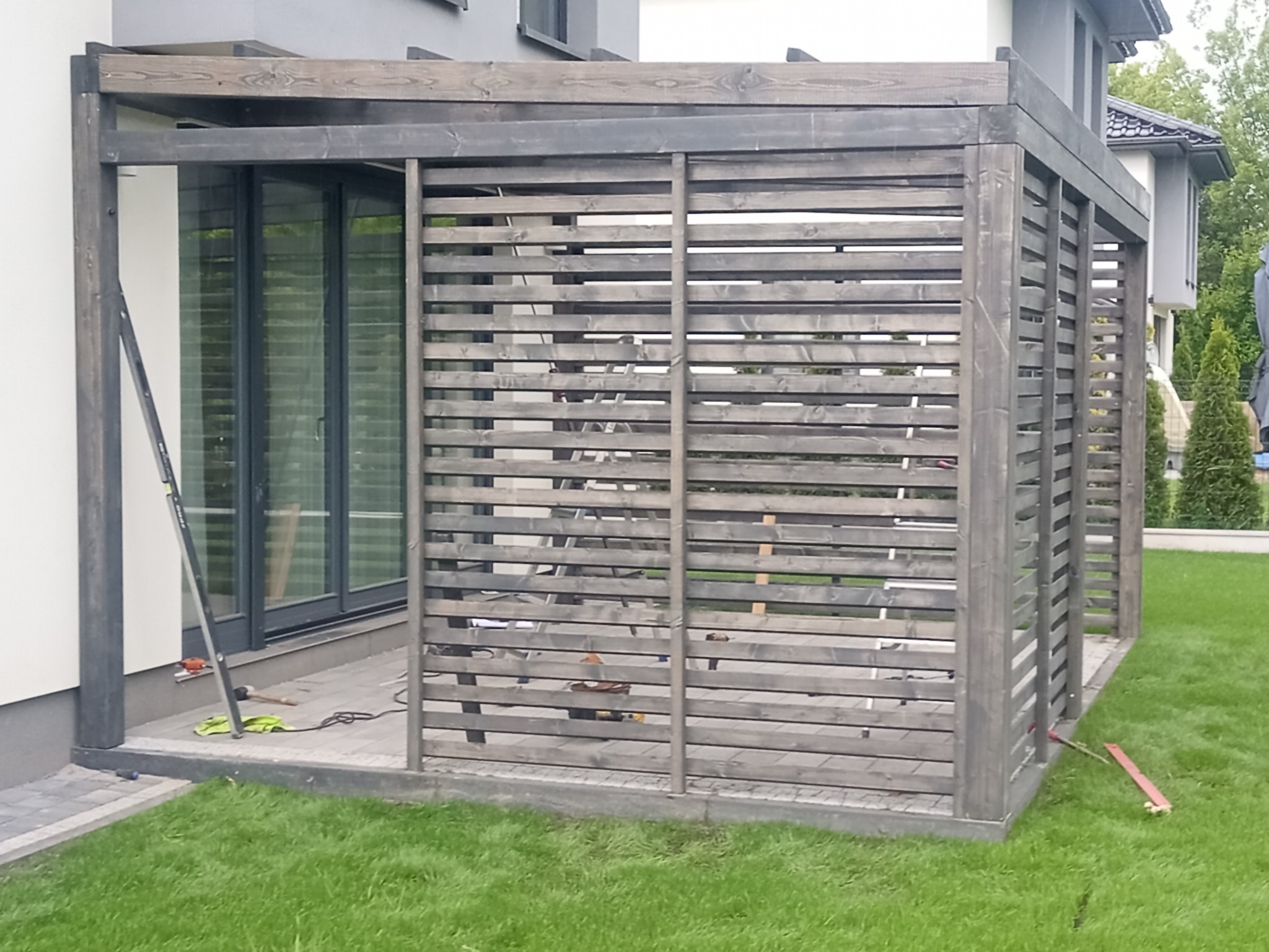 Drewniana pergola w odcieniach szarości z poziomymi listwami, częściowo przysłaniająca okno tarasowe, narzędzia na posadzce, trawnik w tle.