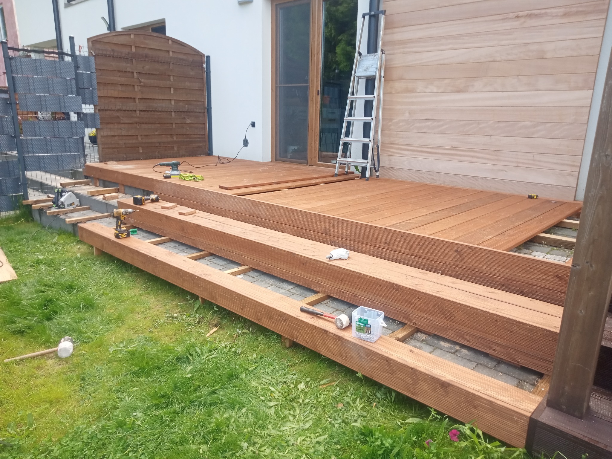 Montaż drewnianego tarasu przy domu. Widoczne deski, legary, narzędzia i drabina. Częściowo ułożona podłoga tarasu. Ujęcie z poziomu trawnika.