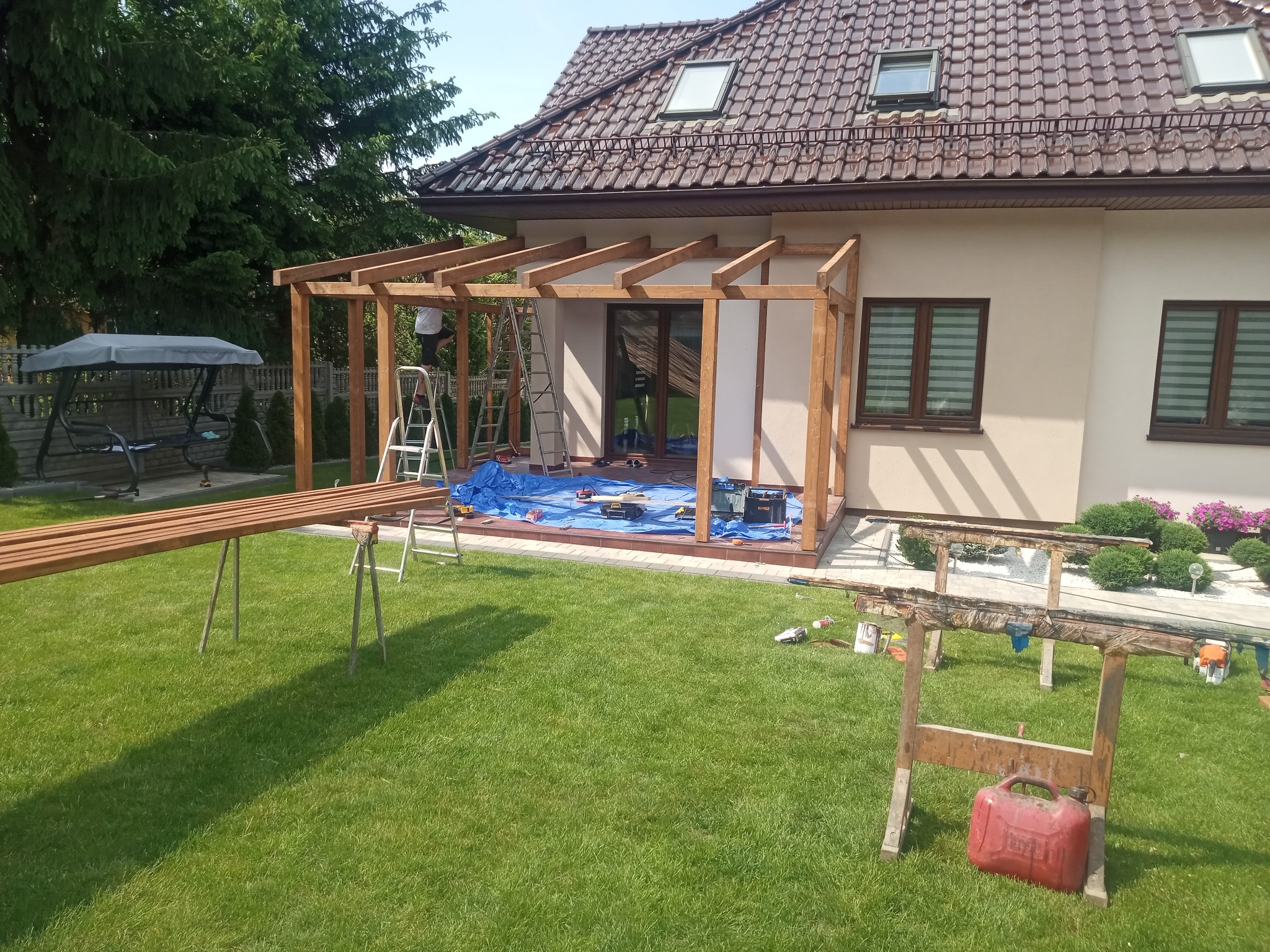 Montaż drewnianej konstrukcji pergoli przy domu jednorodzinnym. Na trawniku leżą narzędzia i materiały budowlane. Prace w toku, widoczny pracownik na drabinie.