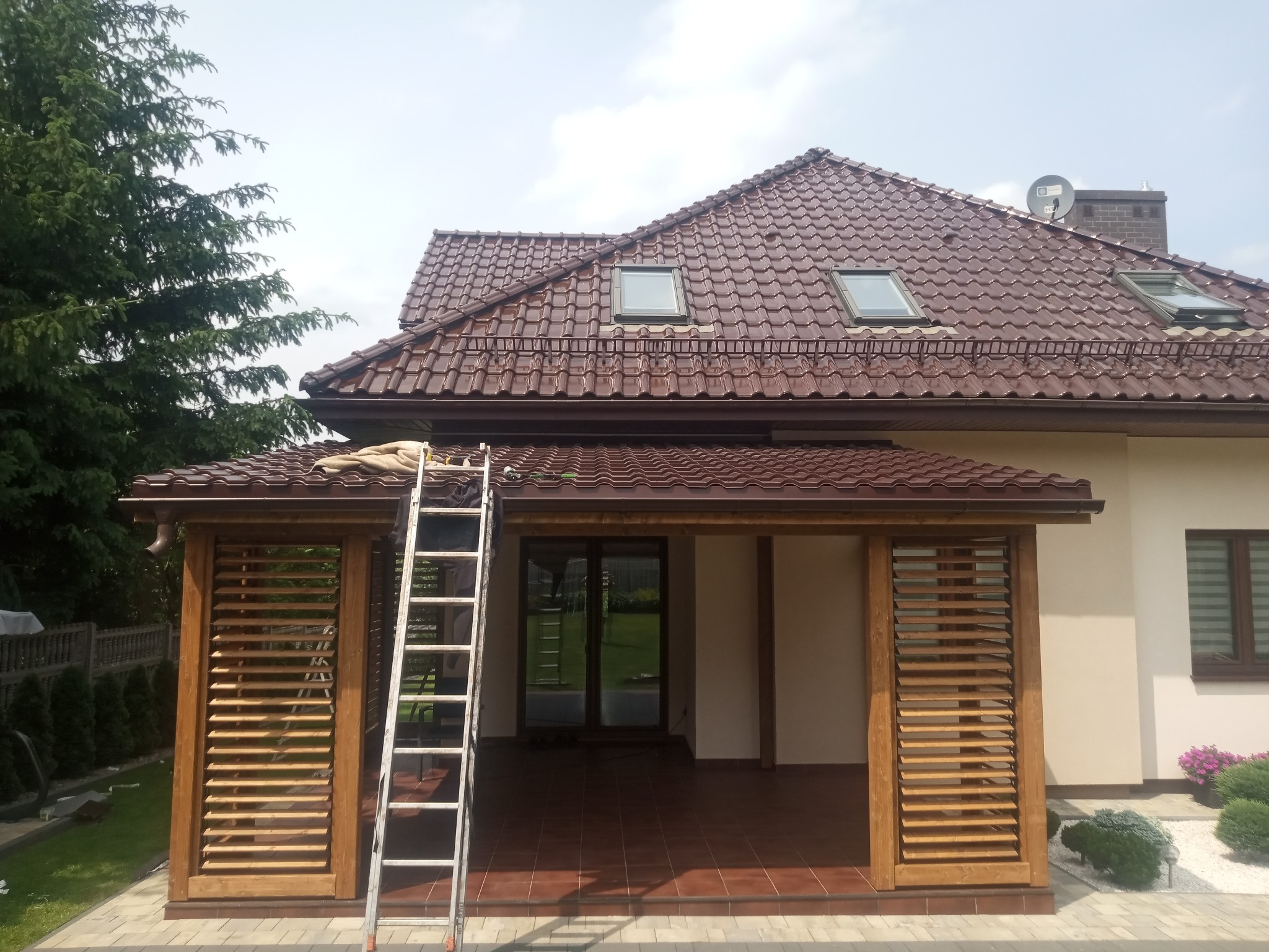 Drewniana pergola z żaluzjami wbudowana w bryłę domu, widoczna drabina, brązowa dachówka, zielone drzewa w tle.