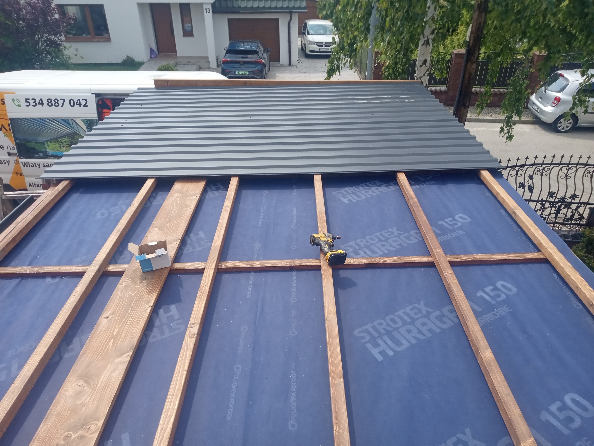 Montaż zadaszenia: drewniany szkielet konstrukcji pokryty folią, częściowo przykryty szarą blachą trapezową. W tle dom z garażem i zaparkowane auta.