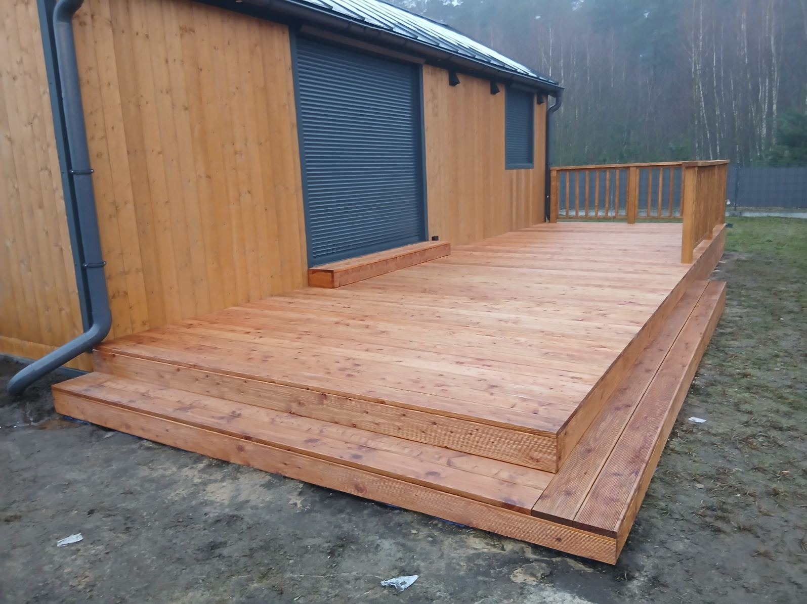 Nowy taras z drewna na zewnątrz budynku z drewnianą elewacją i szarą roletą. Widoczne schody wejściowe i balustrada. Całość w naturalnej kolorystyce drewna.