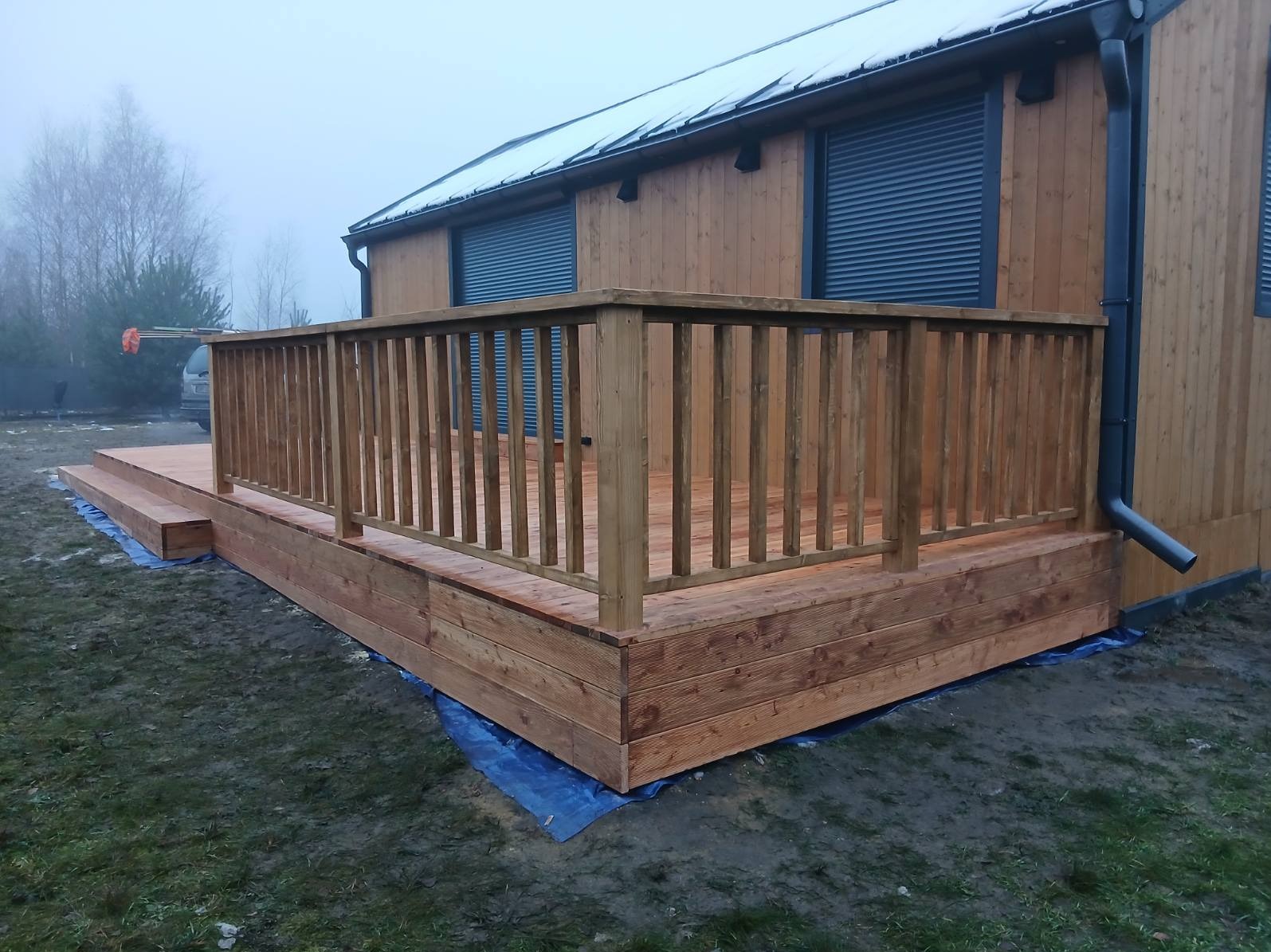 Drewniany taras z balustradą przy domu z drewnianą elewacją i szarymi roletami. Widoczne schody wejściowe. Mglista pogoda.