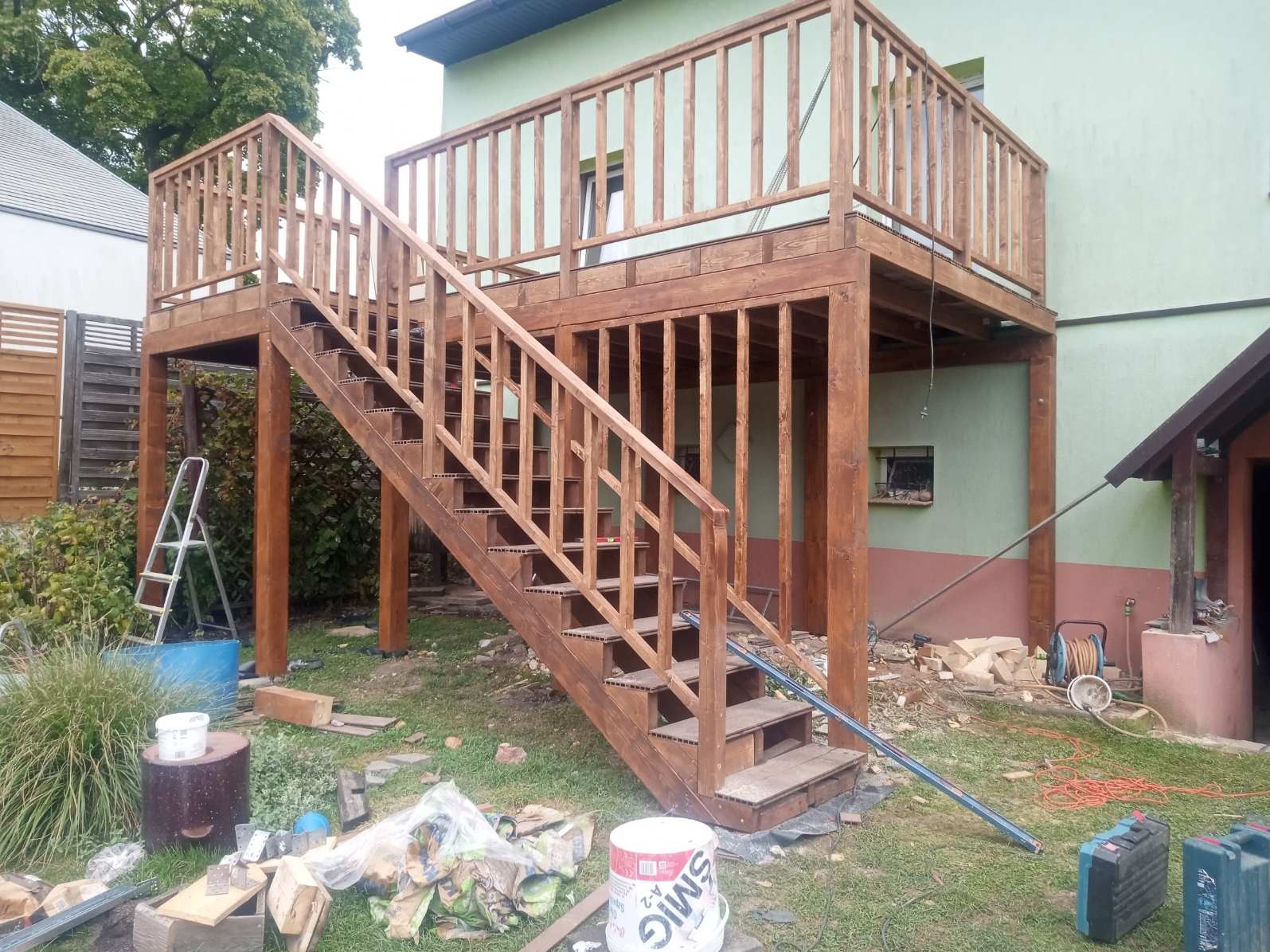 Drewniany taras na podwyższeniu z balustradą i schodami, w trakcie budowy. Widoczne narzędzia i materiały budowlane. Dom z zieloną elewacją w tle.