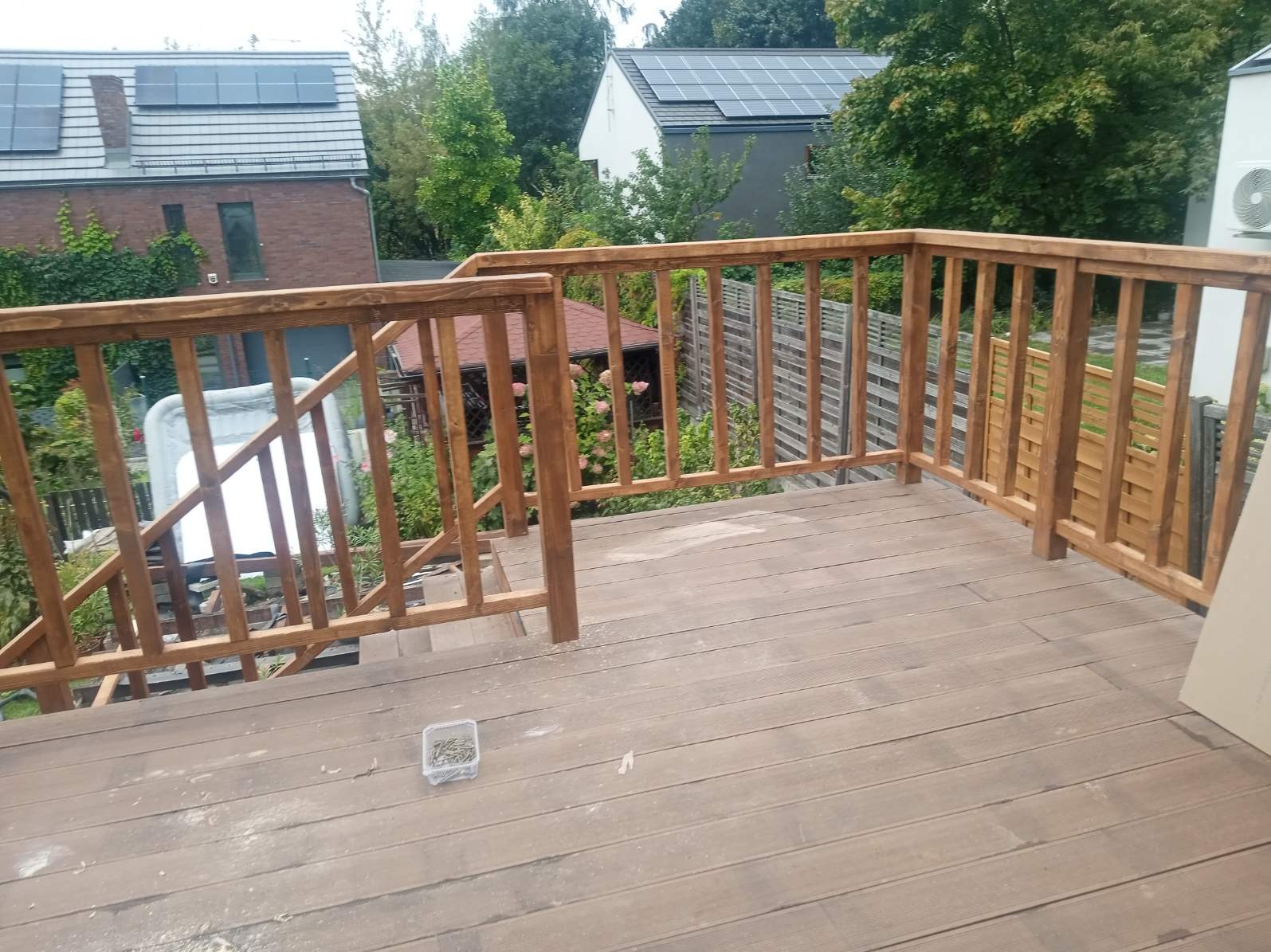 Wykończony taras z brązowych desek kompozytowych z drewnianą balustradą. W tle zieleń i budynki z panelami słonecznymi. Pojemnik ze śrubami na podłodze.