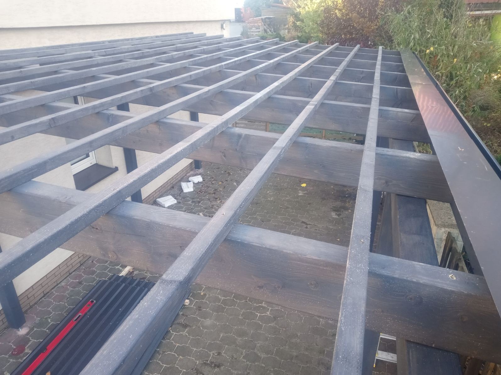 Konstrukcja drewnianej pergoli w trakcie budowy, widok z góry. Szare, pomalowane belki tworzą regularną siatkę nad tarasem z kostki brukowej. Widoczny poziomica.