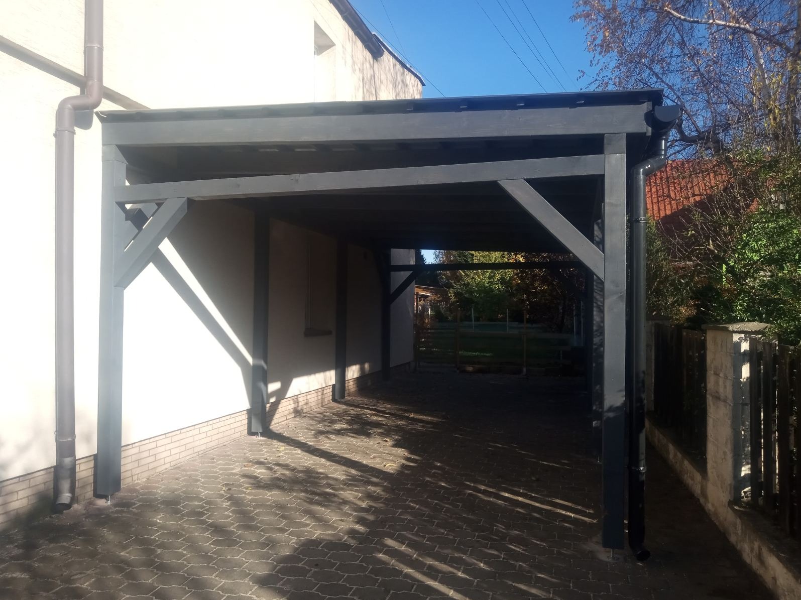 Ciemnoszara, drewniana pergola przy białym budynku, z widocznym brukowanym podjazdem i fragmentem ogrodu w tle. Konstrukcja oświetlona promieniami słońca.