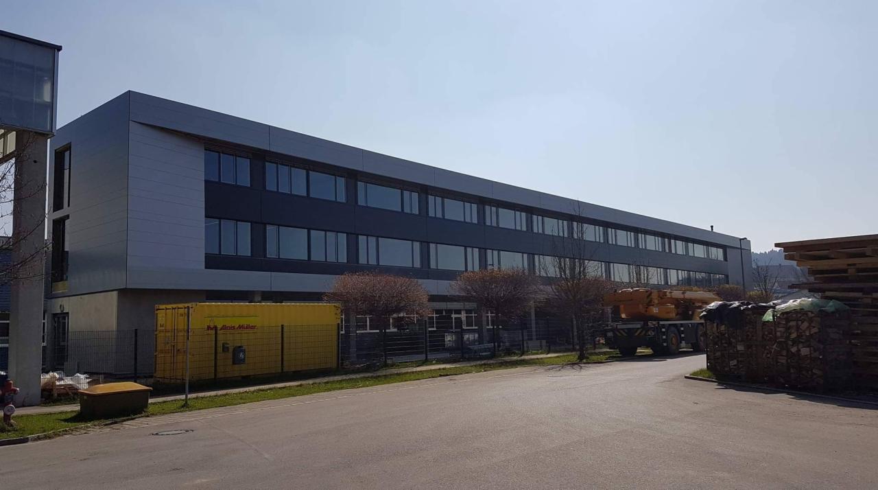 Nowoczesna hala przemysłowa z szarymi panelami elewacyjnymi i rzędem okien, widoczny kontener budowlany Alois Müller i składowane drewno, ujęcie z poziomu ulicy.