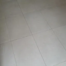 Jasnoszare, matowe płytki podłogowe ułożone w kwadraty, widoczne fragmenty mebli w tle.