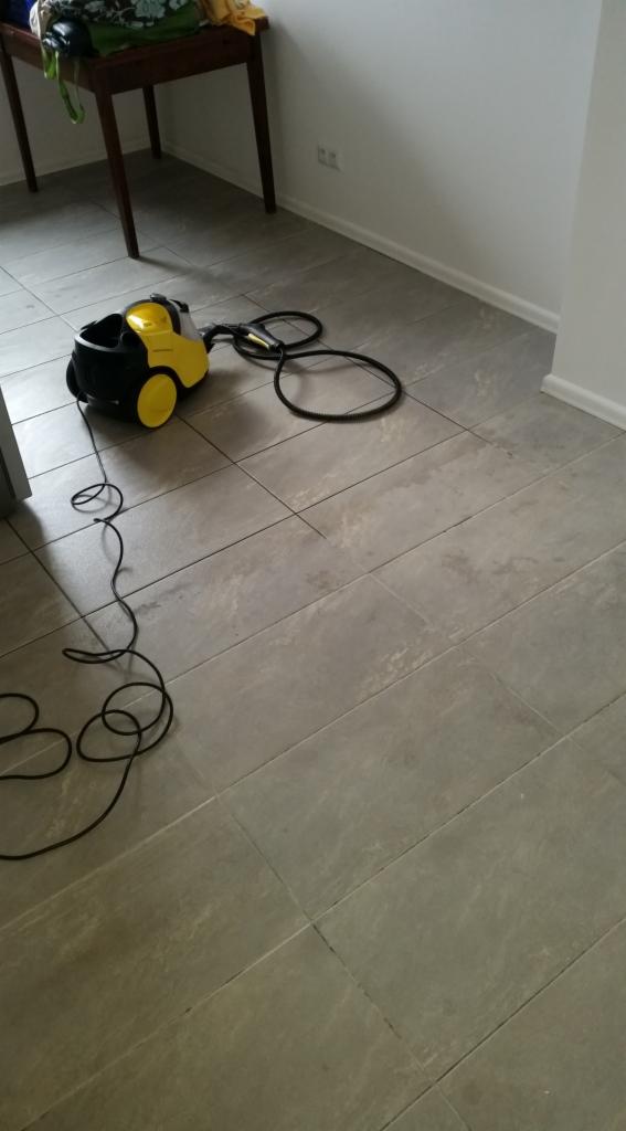 Żółto-czarny odkurzacz przemysłowy na szarych płytkach podłogowych w jasnym pomieszczeniu, z widocznym kablem zasilającym i fragmentem drewnianego stolika w tle.