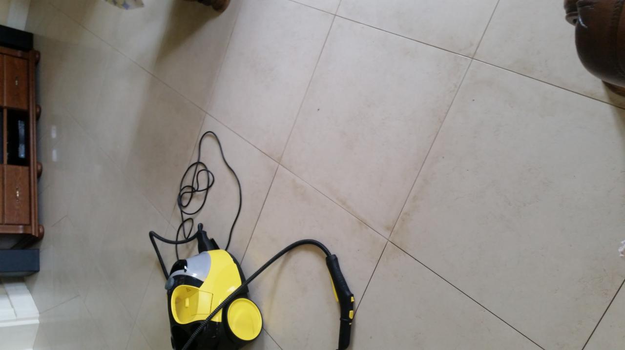 Żółto-czarny parowy mop do podłóg Karcher na jasnych, kwadratowych płytkach podłogowych z widocznymi zabrudzeniami, obok fragmentu mebla.
