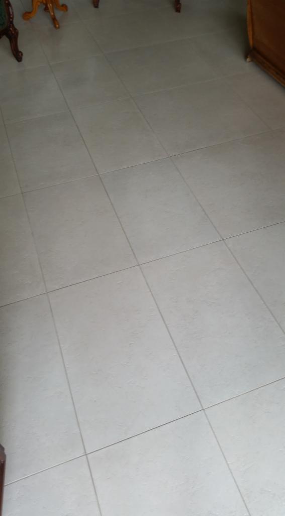 Jasnoszare, matowe płytki podłogowe ułożone w kwadraty, widoczne fragmenty mebli w tle.