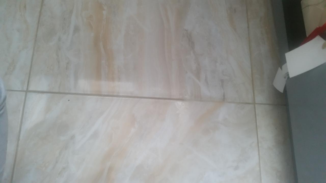 Beżowe płytki podłogowe z delikatnym wzorem marmuru, widoczne fugi, fragment szafki w prawym górnym rogu.